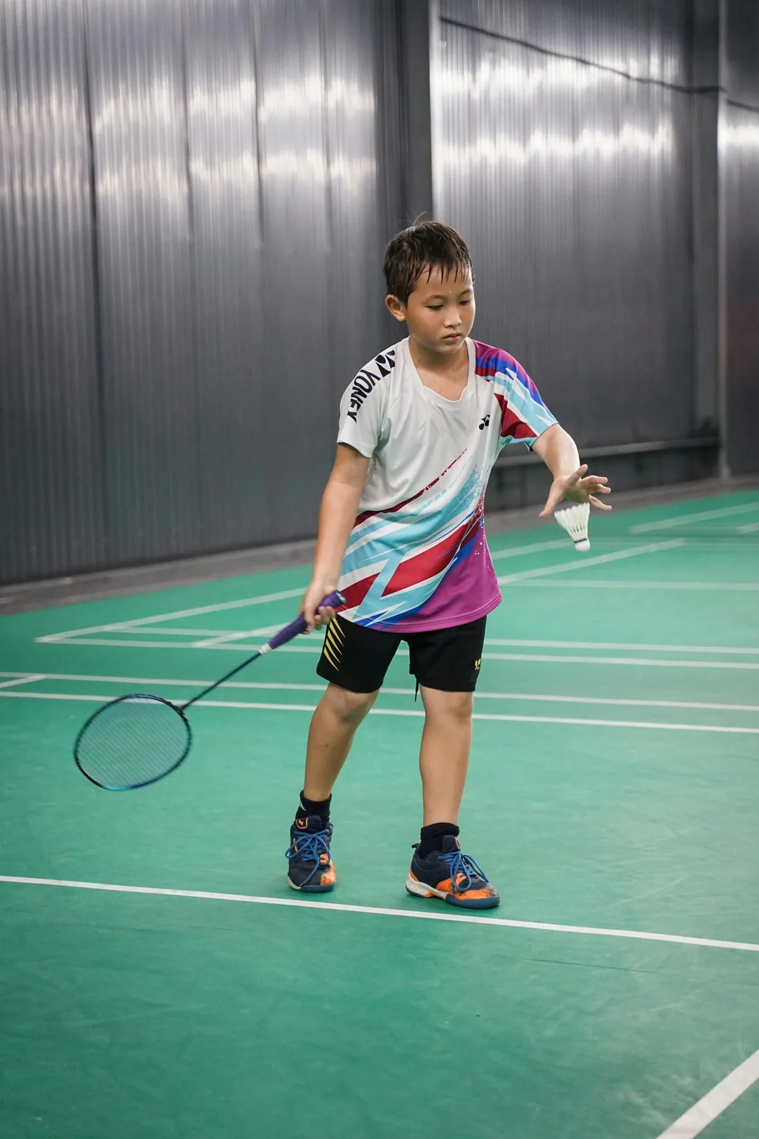 A junior player preparing to serve, shuttlecock in hand, on a quiet indoor court