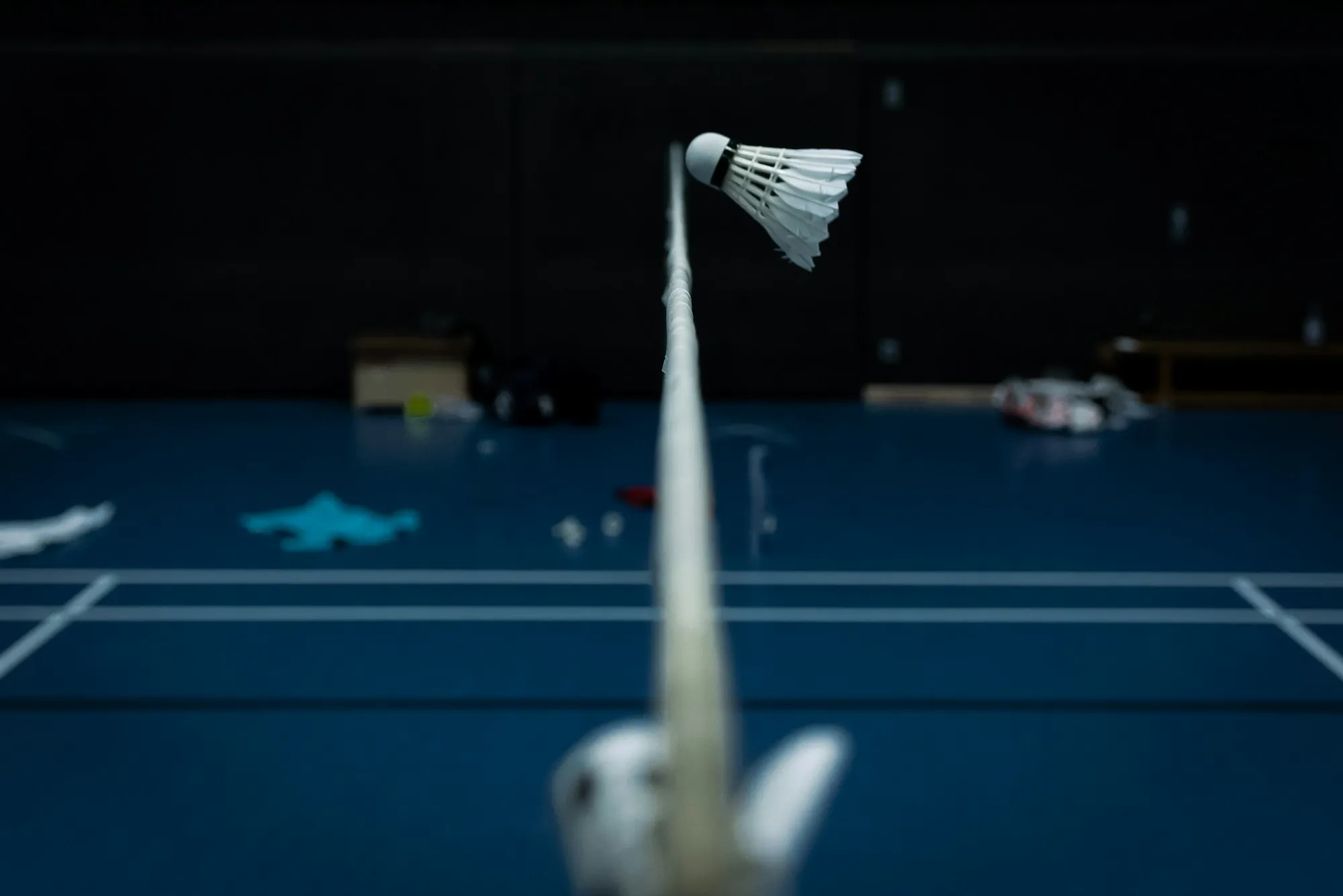 A feather shuttlecock clipping the top of the net tape above a blue indoor court